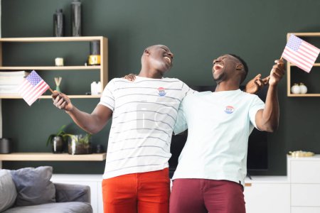 Two young African American men celebrate with American flags, wearing "I Voted" stickers. They are in a home setting, expressing joy and the importance of civic engagement.