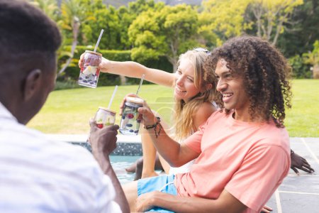 Young Caucasian woman and biracial man toast drinks with an African American man. They are enjoying a sunny outdoor gathering, sharing smiles and casual summer attire.
