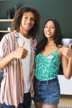 Biracial couple with "I Voted" stickers give thumbs up, smiling confidently. They stand in a home setting, promoting the importance of civic engagement.