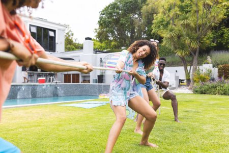 A young biracial woman and a young African American man engage in a playful tug-of-war. They are in a sunny backyard, suggesting a casual outdoor gathering or party.