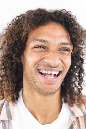 Young biracial man with curly hair laughs joyfully, showcasing a positive mood. His striped shirt adds a casual touch to the bright, upbeat setting.