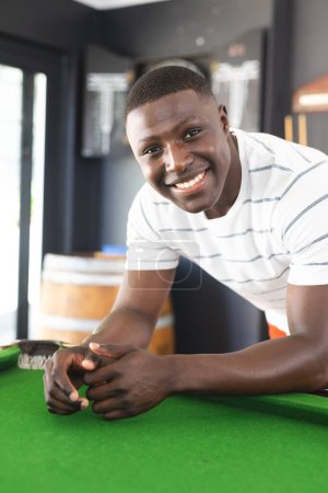 A young African American man smiles while leaning on a pool table. Dressed in a casual white striped polo, he exudes a relaxed and cheerful vibe in a recreational setting.