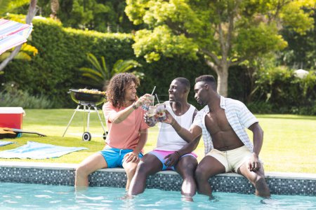 Young African American man and biracial man enjoy drinks by the pool with a friend. Casual summer attire and a barbecue setup suggest a relaxed outdoor gathering.