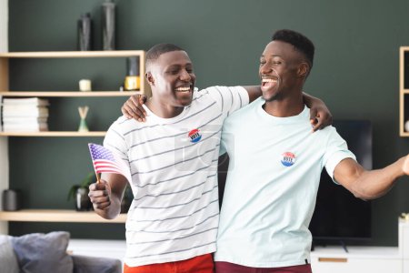 Two young African American men are smiling with "I Voted" stickers, one holding an American flag. They are expressing their civic pride in a home setting, showcasing the importance of voting.