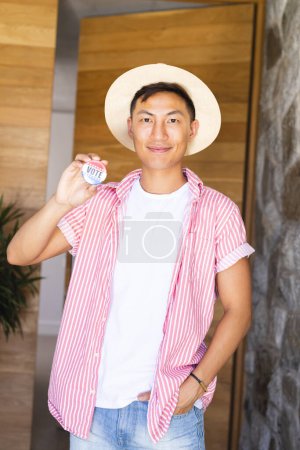 An Asian man holding a 'vote' badge proudly displays it. Captured indoors, his gesture promotes civic engagement and the importance of voting.