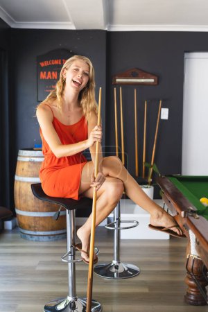 Young Caucasian woman in an orange dress plays pool in a game room. She is seated on a bar stool, laughing and holding a cue stick, exuding a carefree vibe.