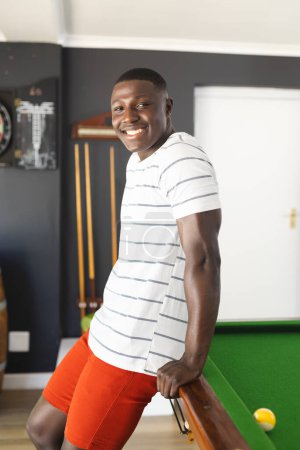 A young African American man leans on a pool table, smiling at the camera. Dressed casually in a striped shirt and red shorts, he exudes a relaxed and happy demeanor in a recreational setting.