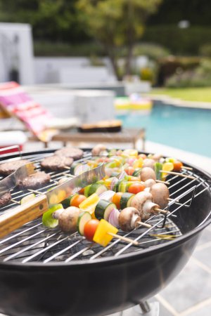 Ein Barbecue-Grill kocht Spieße mit Gemüse und Fleisch am Pool mit Kopierraum. Es wird unter freiem Himmel gekocht, wobei der Schwerpunkt auf einer gemütlichen Mahlzeit im Hinterhof liegt..