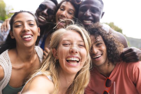 Groupe diversifié d'amis prenant selfie, blotti près avec de larges sourires. Capturées à l'extérieur, leur camaraderie brille à travers leur selfie de groupe spontané.