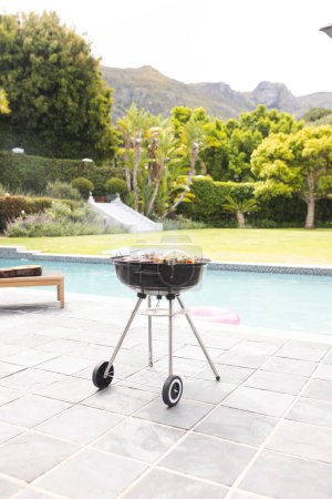 A charcoal grill is ready with food on it, beside a pool in a lush garden with copy space. The setting suggests a leisurely outdoor barbecue in a private backyard with a mountain view.