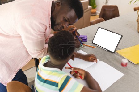 African American father assists a young son with homework at a table with copy space. Father and son engage in educational activities, with a tablet and stationery items present.
