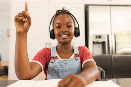 A young African American girl raises her hand during a lesson on a video call online school lesson. She wears headphones, a red shirt, and denim overalls, smiling at her virtual class.
