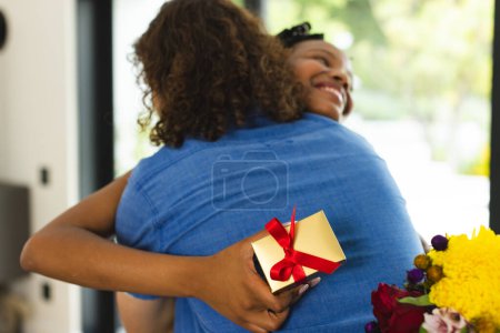African American couple embraces warmly, woman holding gift. She has curly hair, and they're both in casual attire, sharing a joyful moment, unaltered