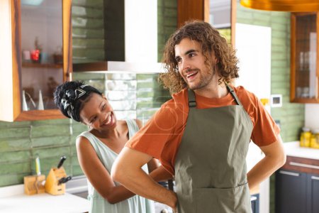 Una pareja diversa atando delantal para cocinar juntos en la cocina. La mujer afroamericana sonríe a su pareja caucásica, que tiene el pelo castaño rizado y una barba, inalterada