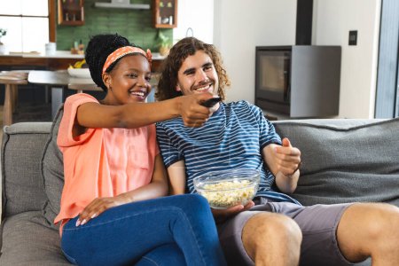 A diverse couple enjoys a cozy moment on the sofa at home, eating popcorn and watching television. The African American woman points at the TV, while her Caucasian partner holds a remote and a bowl of popcorn, both smiling in a relaxed home environme
