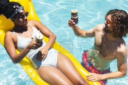 Ein vielseitiges Paar genießt Drinks im Pool. Die Afroamerikanerin mit Kopftuch und der kaukasische Mann, bärtig, stoßen auf Schwebebalken an, unverändert