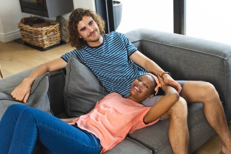 A diverse couple relaxes comfortably on a gray sofa at home. The African American woman has a joyful expression, while the Caucasian man with curly hair smiles gently, unaltered.