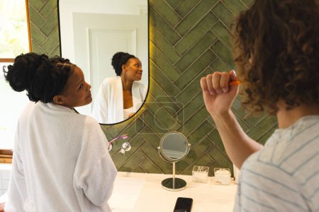 A diverse couple is in well-lit bathroom. The African American woman, with her hair in a bun, watches as her Caucasian partner brushes his teeth, unaltered