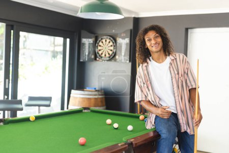 A young African American man with curly hair stands by a pool table with copy space. He is wearing a striped shirt, holding a cue stick, and smiling warmly, unaltered.