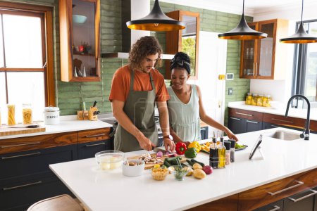 A diverse couple is preparing a meal together in a modern kitchen. The man, with curly hair, and the woman, with her hair in a bun, both wear casual clothing as they chop vegetables, unaltered