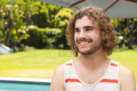 A young Caucasian man smiles warmly near a pool outdoors with copy space at home. He has curly brown hair, light stubble, and is wearing a casual sleeveless top, unaltered.