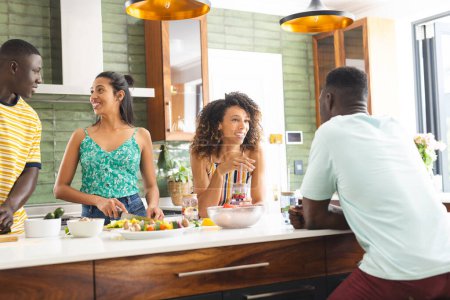 A diverse group of friends gathers in a modern kitchen. The African American male and Hispanic females chat while preparing a meal, showcasing casual attire and lively expressions, unaltered