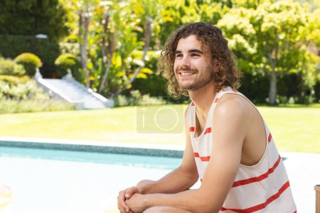 Ein junger kaukasischer Mann mit lockigem braunem Haar lächelt freundlich, mit Kopierraum, zu Hause im Freien. Er trägt ein gestreiftes Tank-Top und sitzt an einem Pool, der eine entspannte, sommerliche Stimmung ausstrahlt, unverändert.