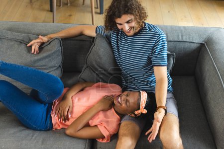 Ein vielseitiges Paar teilt sich einen verspielten Moment auf einer grauen Couch zu Hause. Die Afroamerikanerin mit dem Stirnband lacht und liegt unverändert auf dem Schoß des kaukasischen Mannes..
