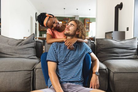 A diverse couple shares tender moment at home. The African American woman, with her hair in a headband, embraces her Caucasian partner from behind, both smiling warmly, unaltered