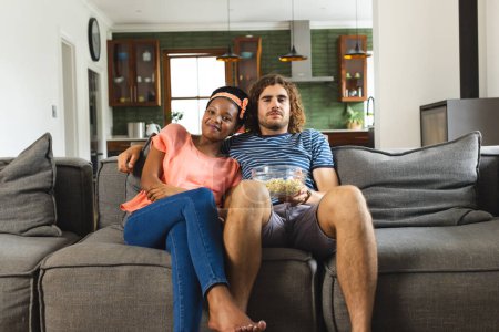A diverse couple is relaxing on a gray sofa at home, eating popcorn and watching television. The African American woman and Caucasian man enjoy a cozy moment, sharing a bowl of popcorn.