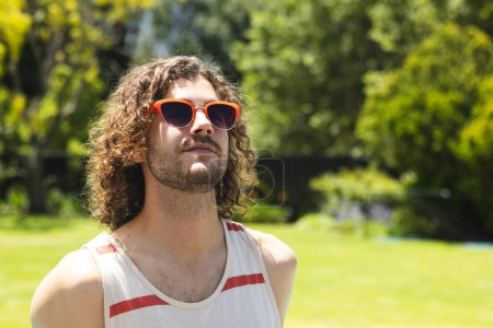 A young Caucasian man gazes upwards, sporting orange sunglasses with copy space at home outdoors. His curly brown hair and casual tank top suggest a relaxed, summery vibe, unaltered.