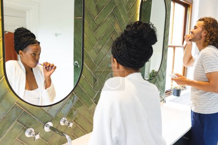 Una pareja diversa está arreglando y cepillando los dientes en un baño bien iluminado. La mujer afroamericana, con el pelo trenzado, se maquilla, mientras que el hombre caucásico se cepilla los dientes, inalterados.