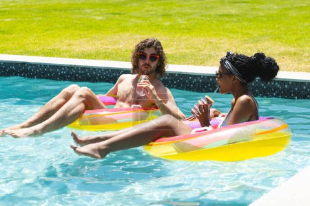A diverse couple relaxes on pool floats, enjoying drinks at home. The Caucasian man and African American woman sport swimwear and sunglasses in sunshine, unaltered