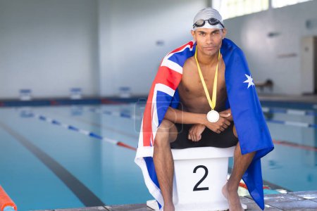 Male athlete swimmer wrapped in Australian flag sitting on podium with gold medal. Wearing goggles and medal, showing pride after swimming competition, unaltered
