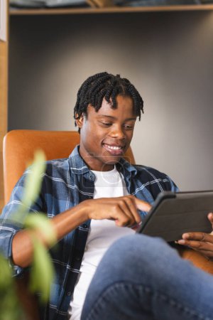 Hombre afroamericano sentado en una moderna oficina de negocios, usando una tableta con una sonrisa. Con ropa casual, tiene el pelo corto y retorcido y un aspecto juvenil, inalterado.
