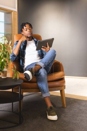 Hombre afroamericano sentado en una moderna oficina de negocios, sosteniendo una tableta, luciendo reflexivo. Con ropa casual, tiene el pelo negro corto y una postura relajada, inalterada.