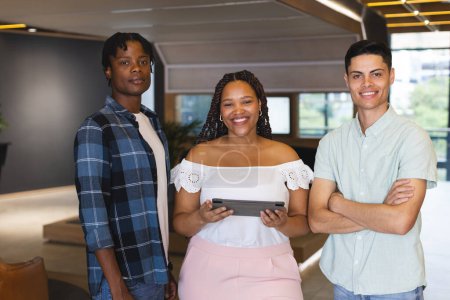 Diverse group of colleagues standing together in a modern business office, one holding tablet. African American man with short hair, biracial woman with braids, and biracial man with neat haircut, all young, smiling, unaltered.