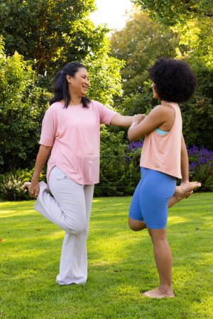 Mature biracial woman helping young biracial woman stretching in garden at home. Both wearing casual clothes, mature woman has long black hair, young woman has curly hair, unaltered