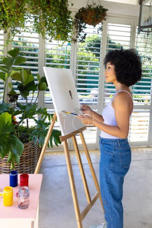 Biracial young woman painting on canvas at home, surrounded by plants. She has curly black hair, wearing casual clothes and holding paintbrush, unaltered