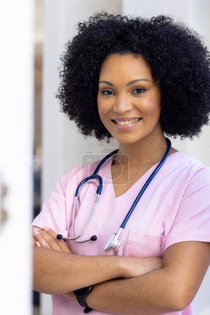 Krankenschwester mit verschränkten Armen, die zu Hause Peelings trägt. Sie hat lockiges schwarzes Haar, hellbraune Haut und lächelt warm, unverändert.