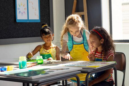 In school, three young girls are focusing on painting in art class. Diverse friends with dark and light brown hair, wearing colorful clothes, are creating art, unaltered.