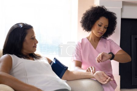Mature biracial woman checking blood pressure at home, young nurse assisting. Both wearing casual clothes, nurse with curly hair and watch, unaltered
