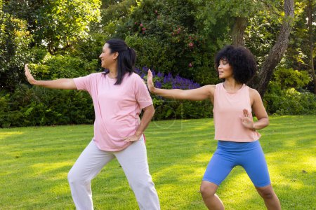 Biracial mother and daughter practicing yoga in garden at home. Mother wearing pink top, daughter in blue, both enjoying exercise, unaltered