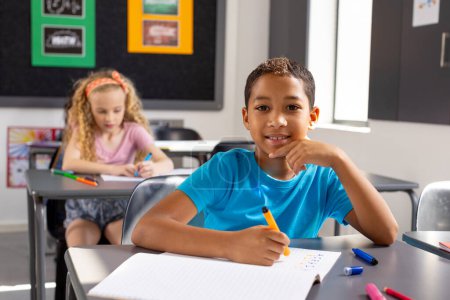 En la escuela, en el aula, un niño birracial y dos niñas caucásicas se centran en su trabajo. Los tres tienen el pelo corto, el chico vistiendo una camisa azul, las chicas con tops estampados, inalterados.