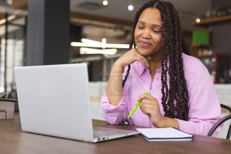 Eine junge Frau mit langen geflochtenen Haaren arbeitet in einem modernen Geschäftsbüro an einem Laptop. Sie trägt ein rosafarbenes Hemd, hält einen Stift in der Hand und hat ein Notizbuch in der Nähe,.