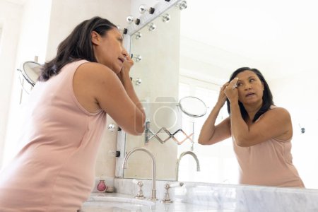 Mature biracial woman removing makeup in the bathroom, looking in mirror at home. She has dark hair, wearing a simple dress, in a bright bathroom, unaltered.