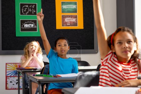 In school, diverse group of young students sitting at desks in a classroom, raising hands.