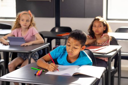 In der Schule, im Klassenzimmer, ein Junge im blauen Hemd, der in ein Notizbuch schreibt, zwei Mädchen schauen zu. Sonnenlicht erfüllt das Klassenzimmer, betont konzentriertes Lernen und neugierige Blicke, unverändert.
