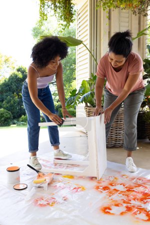 Mère naissante et fille adulte peignent des meubles à l'extérieur à la maison dans le cadre d'un projet d'upcycling. Mère portant une chemise rose a les cheveux noirs courts, fille en bleu a les cheveux bouclés, inchangée.