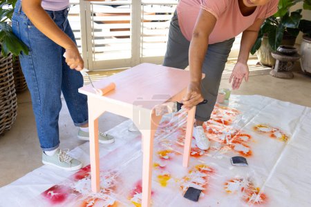 Mature biracial woman painting stool at home in an upcycling project, young biracial woman assisting. Both wearing casual clothes, focused on their DIY project.